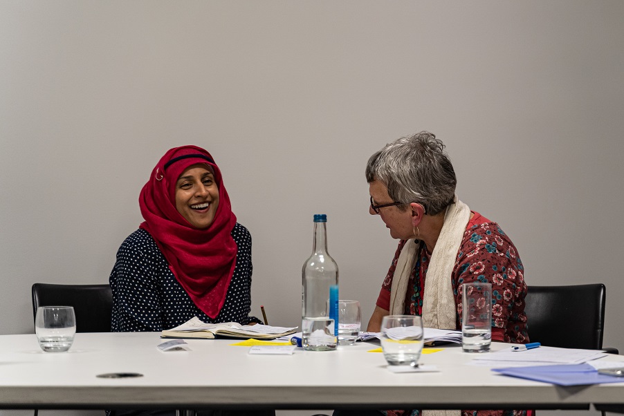 Two women sit together at a table. One is wearing a headscarf, and laughing. One is an older woman with grey hair, looking at the other woman.