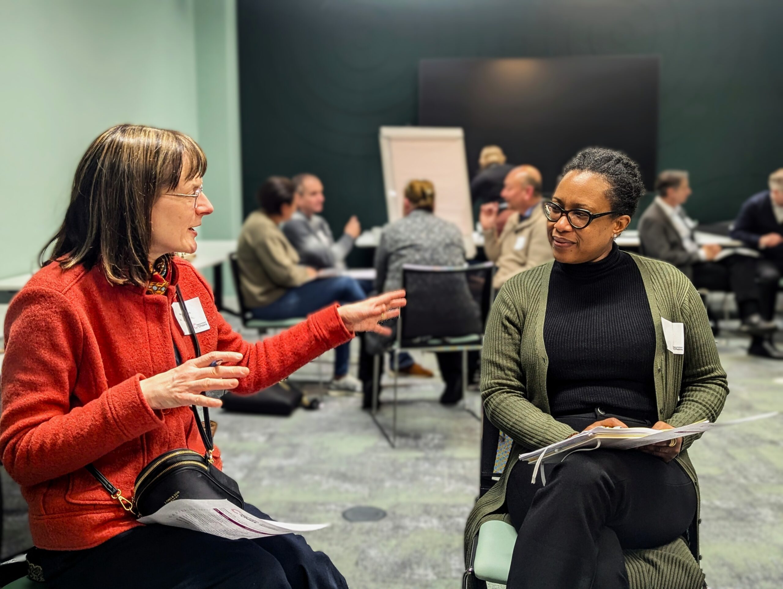 Two charity chairpersons converse at an Association of Chairs event. One gestures while the other listens, holding papers.