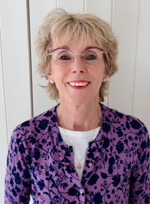 Photo of Roma Hooper - a woman with short blonde hair and glasses, wearing a purple cardigan with black flowers. She is looking at the camera and smiling.