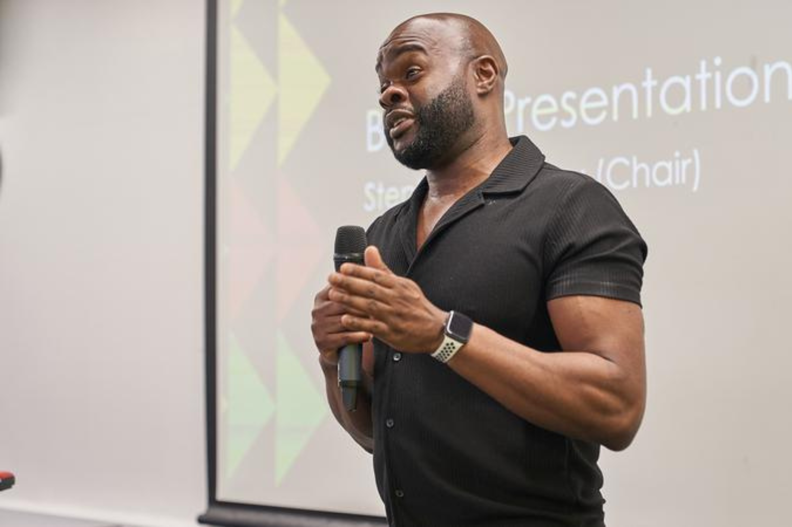 A photo of Stephen Bunbury, a Black man wearing a black polo shirt, is standing in front of a projection screen. His is holding a microphone and talking.